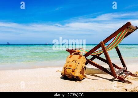 A deck chair on the sandy shore of a tropical beach overlooking the open sea. a stylish brown backpack standing nearby. A place for the traveler, free Stock Photo