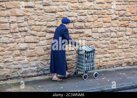 An elderly lady wearing a coat and beret, pushes her walker or shopping trolley past a limestone wall, Dorset, England. Stock Photo