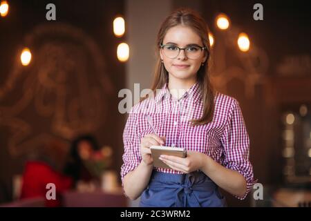 Portrait of a beautiful friendly waitress in glasses with notepapers and pen ready to take your order Stock Photo