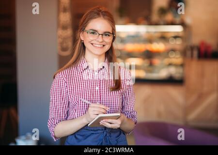 Portrait of a beautiful friendly waitress in glasses with notepapers and pen ready to take your order Stock Photo