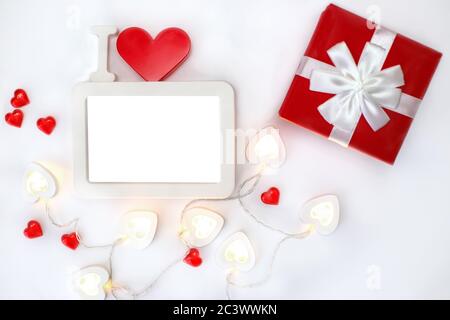 A white empty rectangular photo frame with a red heart and a red box with a white bow lies on a white background. Nearby lie bright luminous garlands Stock Photo