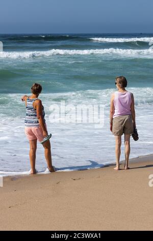 Two relaxed looking senior woman on beach walking at edge of sea with white waves rolling. Portugal Stock Photo