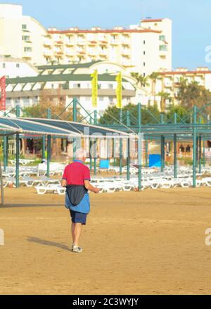 Side, Turkey - April 12 - 2019: An elderly Caucasian man in a red T-shirt and shorts is doing exercises on an empty beach. The view from the back. Stock Photo