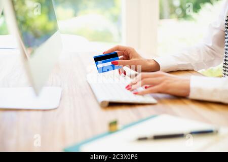 Close-up of woman’s hand holding bank card and typing on keyboard while sitting at desk and shopping online. Focus on bank card. Stock Photo