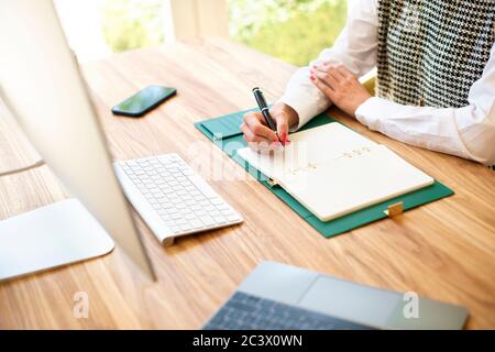 Shot of unrecognizable woman sitting at desk in front of her computer and writing something into the notebook. Stock Photo