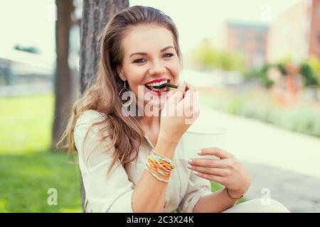 Young positive Latina woman eating delicious salad in park, laughing Stock Photo