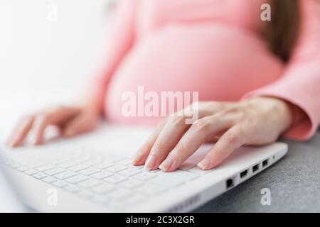 Close up of pregnant woman working on laptop while sitting on bed at home. Stock Photo