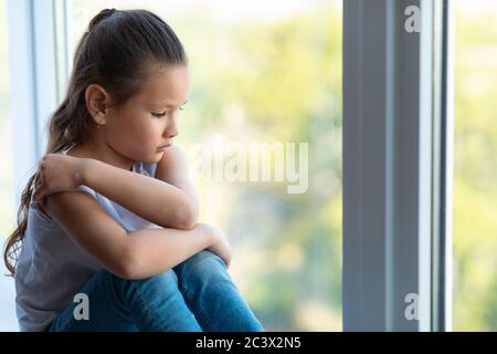 Unhappy Kid Girl Sitting Near Window Suffering From Loneliness Indoors Stock Photo