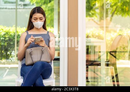 Asian woman customer wear face mask using her mobile phone while sit in waitting social distance queue in front of restaurant with free copyspace for Stock Photo