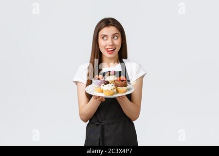 Grocery store employees, small business and coffee shops concept. Excited cute female barista in black apron holding plate with delicious cupcakes and Stock Photo