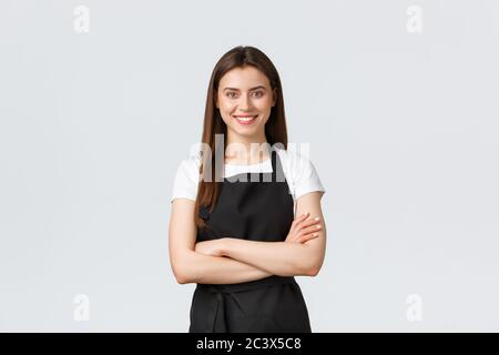 Cheerful smiling female barista in black apron cross arms chest, looking ready and confident. Young girl employee open coffee shop, greeting customers Stock Photo