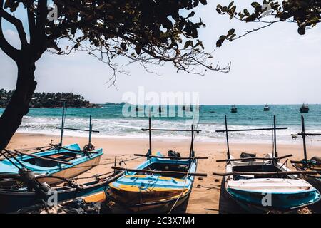 Idle fishing boats tied to the shore at a secluded beach in Sri Lanka. Blue ocean, windy weather and waves in the background. Summer remote vacation Stock Photo