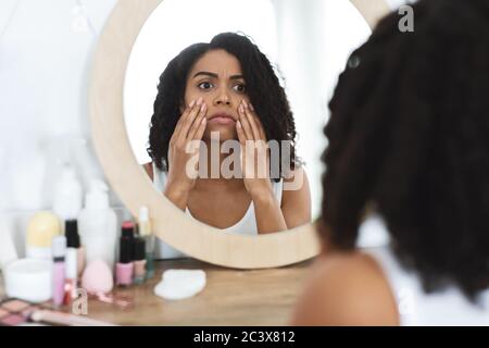 Anti-Wrinkle Skincare. Upset Black Woman Touching Her Face, Looking In Mirror Stock Photo