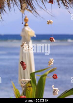 Red and white coral pieces decorations design, hanging on wire at exotic beach bar, with blurred umbrella and sea waves in background, on Gili Air isl Stock Photo