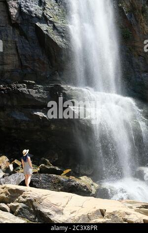 Blue haired girl in a hat staying under Ramboda waterfall in Sri Lanka. Watching water flowing at a popular touristic viewpoint in Ella. Stock Photo