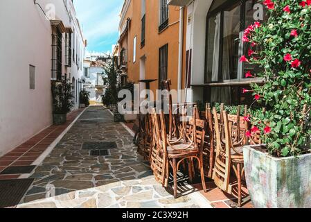 Outside view of a closed cafe during early morning in Spain. Bright sun making shadows on stacked chairs and colorful yellow and white buildings. Stock Photo