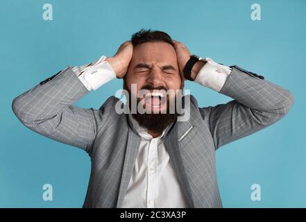 Millennial businessman holding his head and screaming in anger on blue background Stock Photo