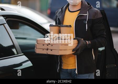Courier in protective mask and medical gloves take order from car. Delivery boy hold pizza and coffe. Delivery service under quarantine. Coronavirus Stock Photo