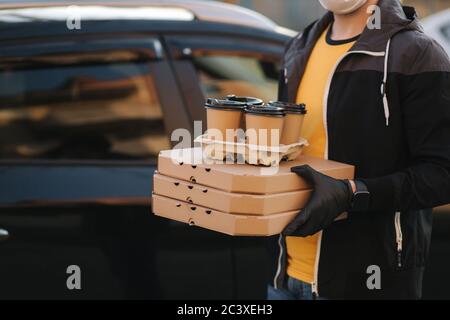 Courier in protective mask and medical gloves take order from car. Delivery boy hold pizza and coffe. Delivery service under quarantine. Coronavirus Stock Photo