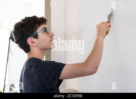 Masonry work in the apartment: a Caucasian boy is intent on filling the imperfections of the wall. On the scraper there is a dose of putty paste that Stock Photo
