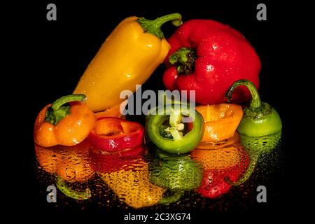 Brightly Colored Peppers and pepper slices covered in water drops on a wet reflective surface against a black field ready for preparation. Plenty of r Stock Photo