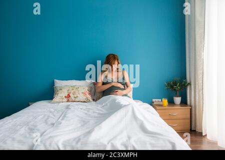 A smiling pregnant woman in a semi-sitting position in bed with her hands around her stomach. She's looking down at her stomach. Stock Photo