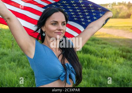 Woman wearing jeans and blue top holding an American flag above her head on a nature during sunset celebration. Stock Photo