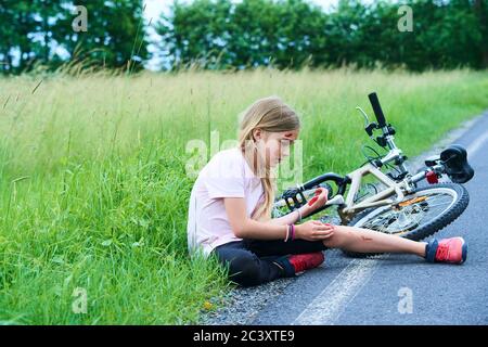 Sad crying little child girl fell from the bike in the summer park. Bleeding on hands and feet. Bicycle accident. Injuries while cycling. Stock Photo