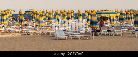 Rows of folded, blue-and-yellow striped parasols and white sunbeds line the beach of Albena on the Black Sea, Balchik Municipality, Bulgaria, in the evening light. Stock Photo