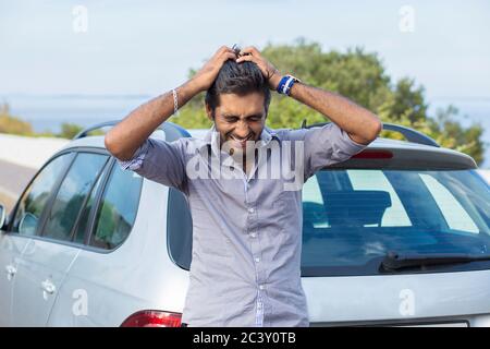 Upset driver after car accident crying hands on head, or he forgot the key while standing outdoors near his car, green tree blue sea and sky on backgr Stock Photo