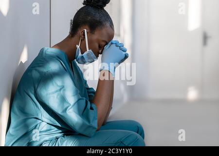 Tired depressed female african nurse wearing face mask sits on hospital floor. Stock Photo