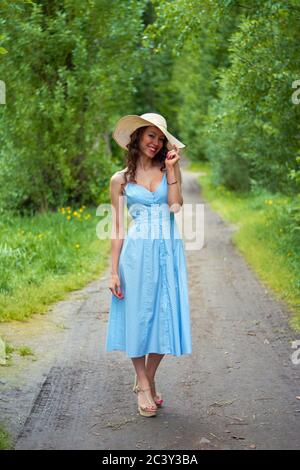 beautiful girl in a straw hat and blue dress on the road in the Park.curly brunette with a smile on her face
