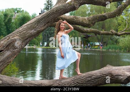 a beautiful curly haired girl in a blue dress stands on a large tree against the background of a Park with a lake Stock Photo