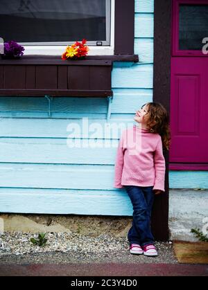 A young girl stands in front of a door and looks up at potted flowers.