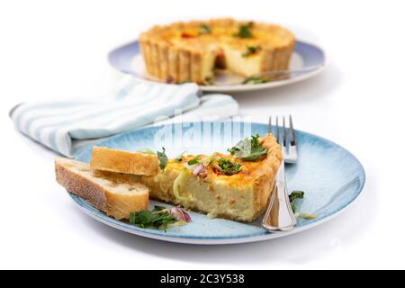 a slice of a cheese quiche on a small plate with slices of French bread with the whole pie out of focus in the background isolated on white Stock Photo