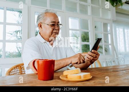 Happy elderly Asian man smiling and using mobile phone while sitting at table with hot drink and bread in morning at home Stock Photo