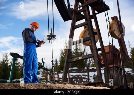 Oil engineer, wearing blue overalls and orange helmet, with a laptop standing next to an oil pump jack, making notes in his computer, low angle view Stock Photo