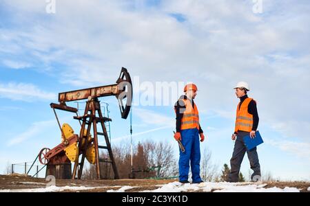 Two oil men in helmets and work vests standing near oil well pump jack and discussing work. Oil worker holding clipboard and talking with colleague at oil field. Concept of petroleum industry. Stock Photo