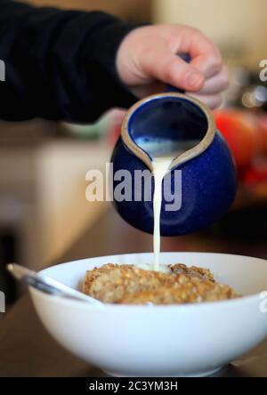 hand pouring milk from small blue jug into cereal bowl, selective focus Stock Photo