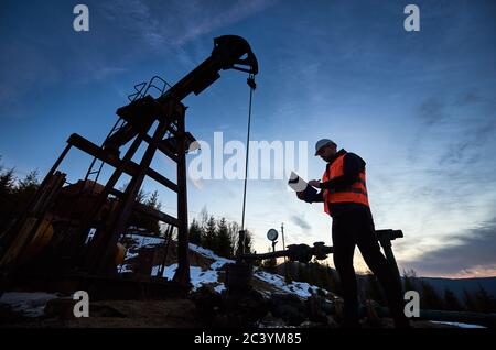 Low angle of petroleum engineer in work vest looking at oil pump rocker-machine and writing on clipboard. Oil worker checking work of balanced beam petroleum pump jack under beautiful evening sky. Stock Photo