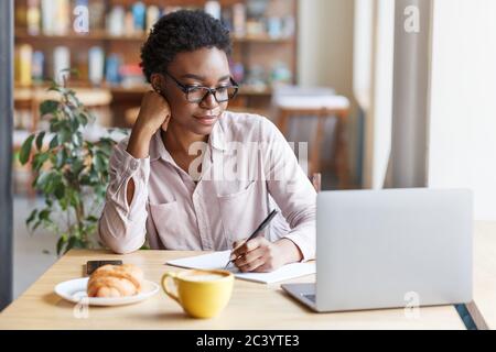Online education. Serious black girl studying remotely on laptop from city cafe Stock Photo