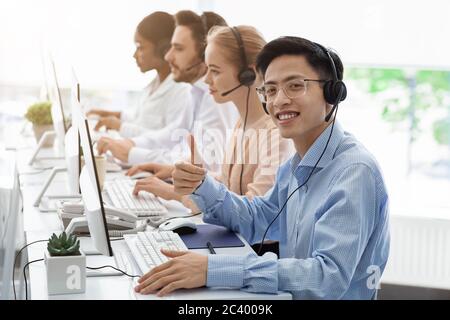 Call center worker showing thumb up gesture and his team with headphones working in open space office Stock Photo