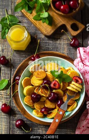 Tiny pancakes breakfast. Mini pancakes with cherries and honey on rustic wooden table. Stock Photo