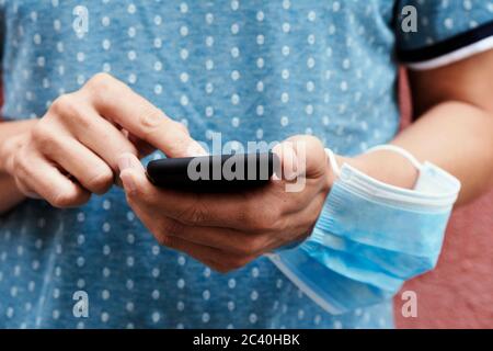 closeup of a young man on the street, in casual wear, using his smartphone and wearing his surgical mask in his arm while is not wearing it in his fac Stock Photo