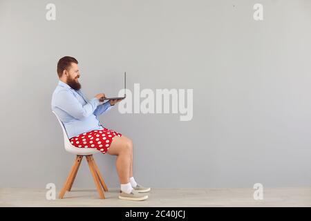 Funny fat man in a blue shirt and red shorts sitting on a chair working online using a laptop on a gray background. Stock Photo