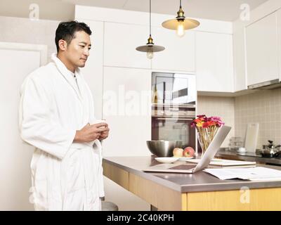 young asian business man in bathrobe working from home standing by kitchen counter holding a cup of coffee looking at laptop computer Stock Photo