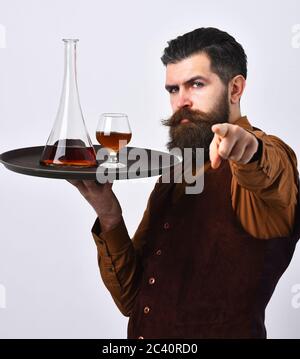 Barman with serious face serves scotch or brandy. Waiter with glass and bottle of whiskey on tray points at you. Man with beard holds cognac on white background. Expensive beverages concept. Stock Photo