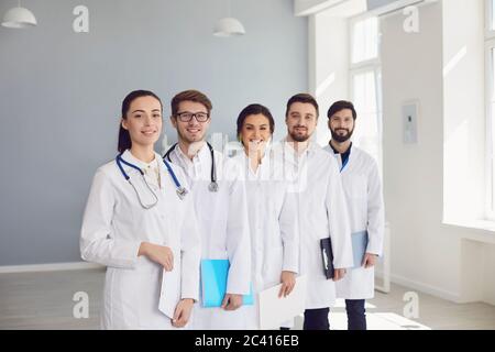 A group of confident practicing doctors in white coats are smiling against the backdrop of the clinic. Stock Photo