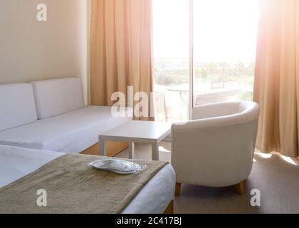 Sunny room in the hotel in light colors with a bed, white table, sofa and armchair. New white disposable slippers in a transparent package lie on the Stock Photo