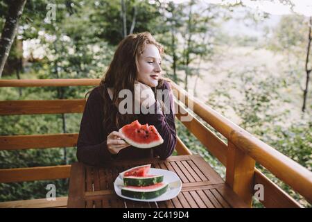 Young redhead woman eating watermelon on wooden house balcony Stock Photo